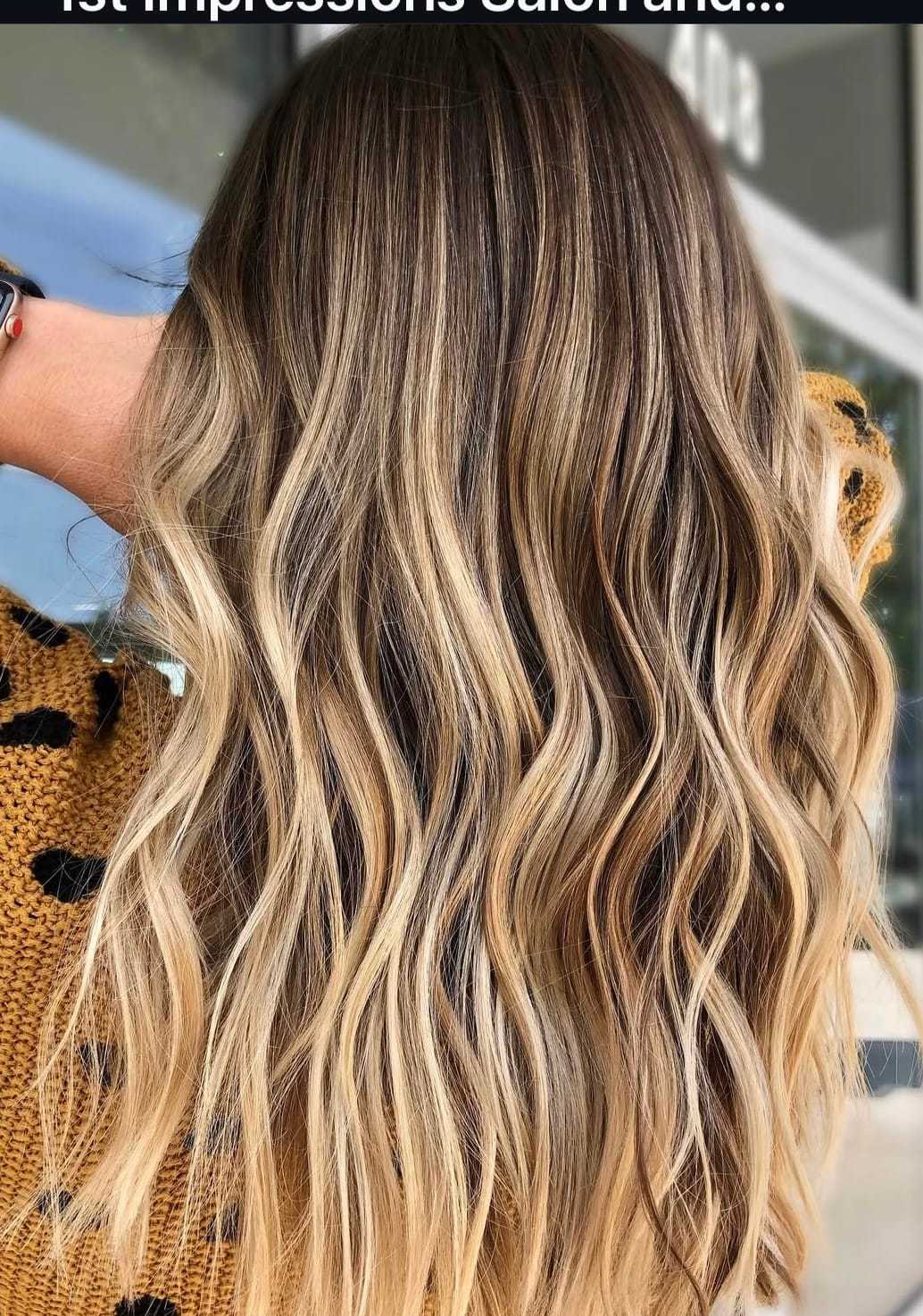 Wavy blonde balayage hair on a person wearing a patterned sweater against a blurred background.