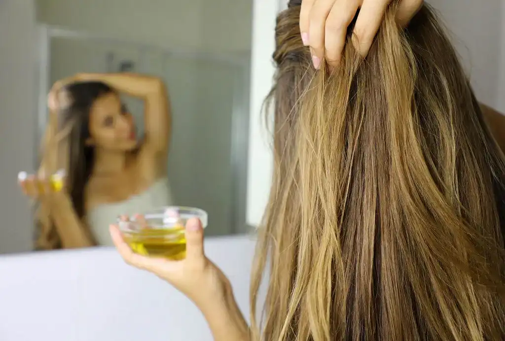 Woman applying hair oil while looking in a bathroom mirror.